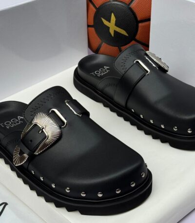 A pair of black leather Toga Pulla mule slides with silver buckles and studs, presented in a white box with the brand's logo.