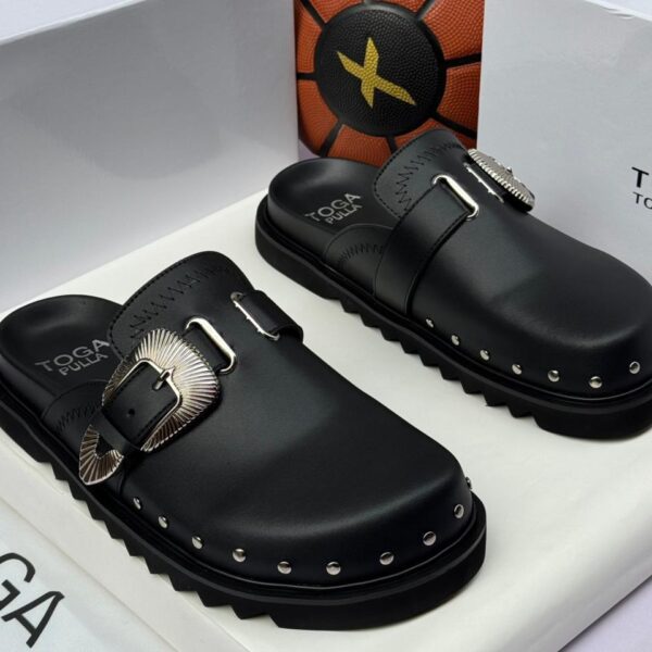 A pair of black leather Toga Pulla mule slides with silver buckles and studs, presented in a white box with the brand's logo.