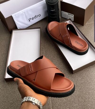 A pair of brown double strap slide sandals with black sole, presented in an open box with a luxurious setting.
