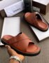 A pair of brown double strap slide sandals with black sole, presented in an open box with a luxurious setting.