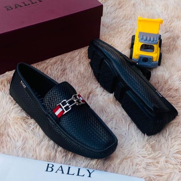 A pair of black leather loafers with a red and white strap on a plush beige surface, accompanied by a maroon Bally box and a yellow toy truck.