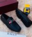 A pair of black leather loafers with a red and white strap on a plush beige surface, accompanied by a maroon Bally box and a yellow toy truck.