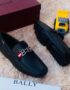 A pair of black leather loafers with a red and white strap on a plush beige surface, accompanied by a maroon Bally box and a yellow toy truck.