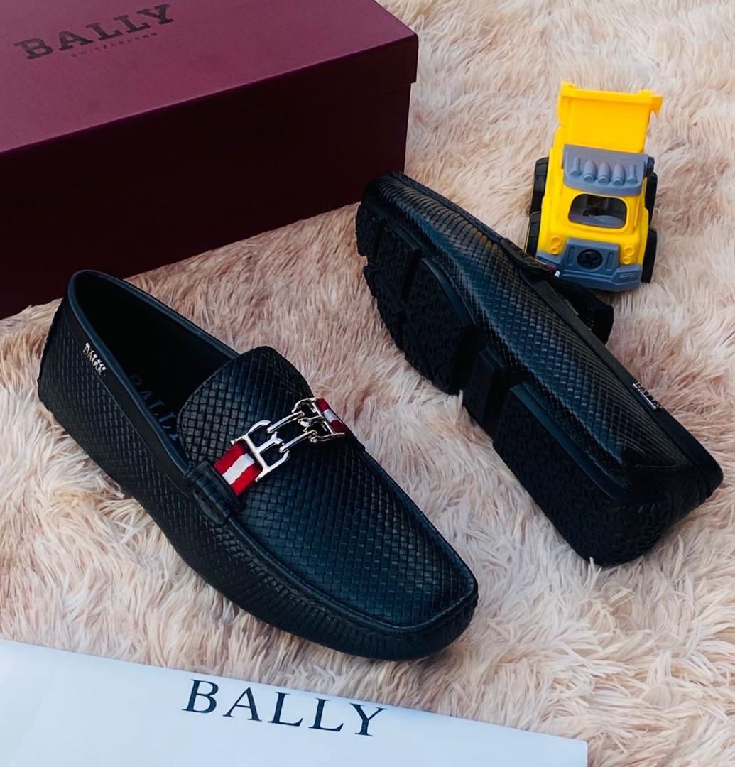 IMG-20260425-WA0009 A pair of black leather loafers with a red and white strap on a plush beige surface, accompanied by a maroon Bally box and a yellow toy truck.