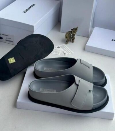 A pair of gray leather Jil Sander slide sandals with black outsole, displayed on a white box with the brand's logo and packaging in the background.