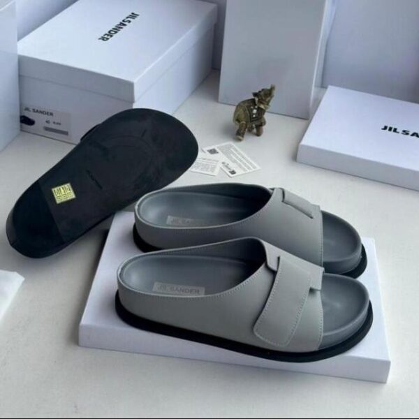 A pair of gray leather Jil Sander slide sandals with black outsole, displayed on a white box with the brand's logo and packaging in the background.