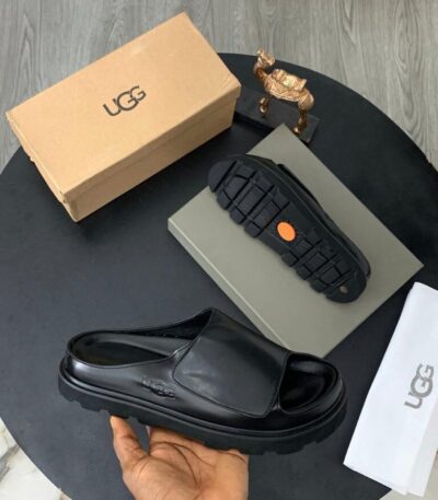 A pair of black UGG leather slide sandals with original box, dust bag, and packaging on a stylish black table.