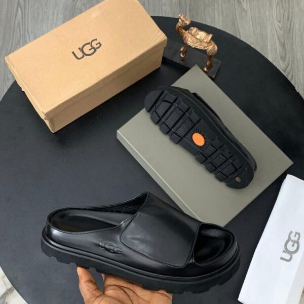 A pair of black UGG leather slide sandals with original box, dust bag, and packaging on a stylish black table.