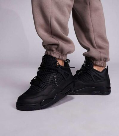 A pair of black high-top sneakers with perforated details on a person's feet, dressed in beige sweatpants, against a light grey background.