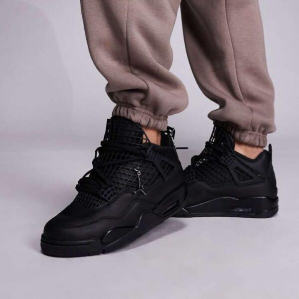 A pair of black high-top sneakers with perforated details on a person's feet, dressed in beige sweatpants, against a light grey background.