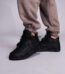 A pair of black high-top sneakers with perforated details on a person's feet, dressed in beige sweatpants, against a light grey background.