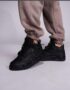 A pair of black high-top sneakers with perforated details on a person's feet, dressed in beige sweatpants, against a light grey background.