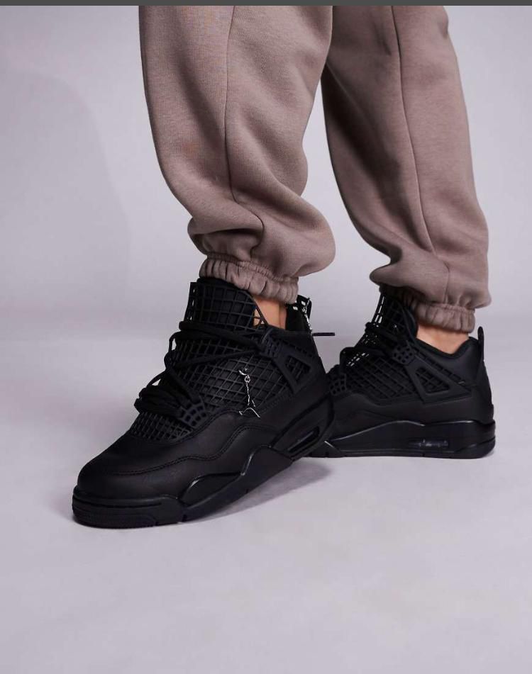 IMG-20260425-WA0017 A pair of black high-top sneakers with perforated details on a person's feet, dressed in beige sweatpants, against a light grey background.