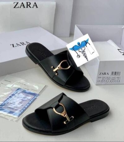 A pair of black leather slide sandals with a gold buckle, displayed with their white box and packaging on a clean white surface.
