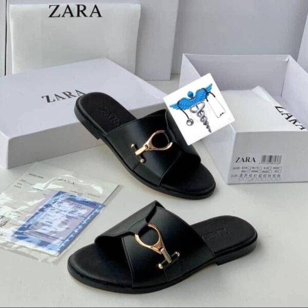 A pair of black leather slide sandals with a gold buckle, displayed with their white box and packaging on a clean white surface.