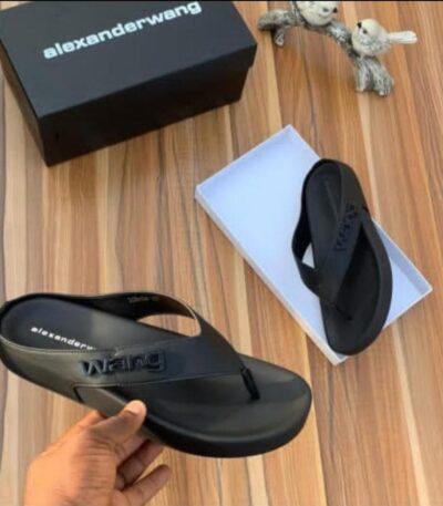 A pair of black rubber thong sandals with an embossed Alexander Wang logo, displayed on a wooden surface with the brand's box and packaging.