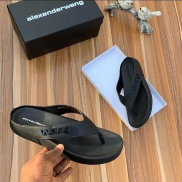 A pair of black rubber thong sandals with an embossed Alexander Wang logo, displayed on a wooden surface with the brand's box and packaging.