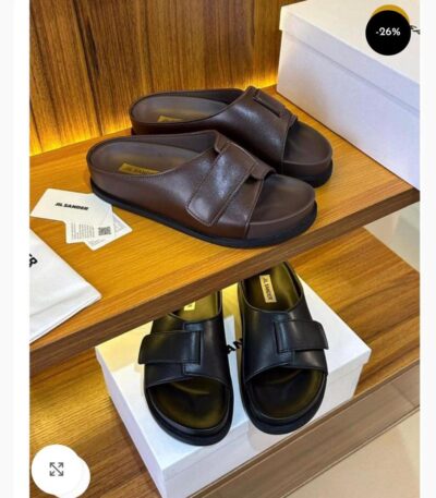 A pair of brown and black Jil Sander double strap leather slides displayed on a wooden shelf with their respective boxes and packaging.