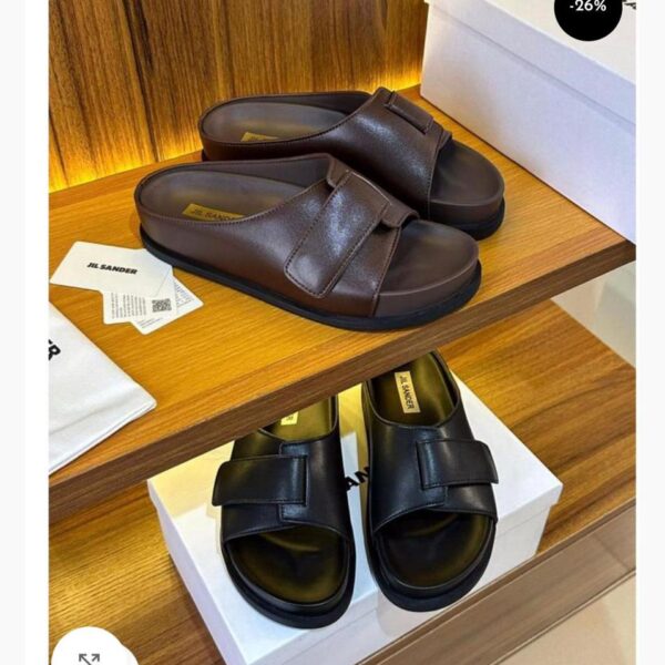 A pair of brown and black Jil Sander double strap leather slides displayed on a wooden shelf with their respective boxes and packaging.