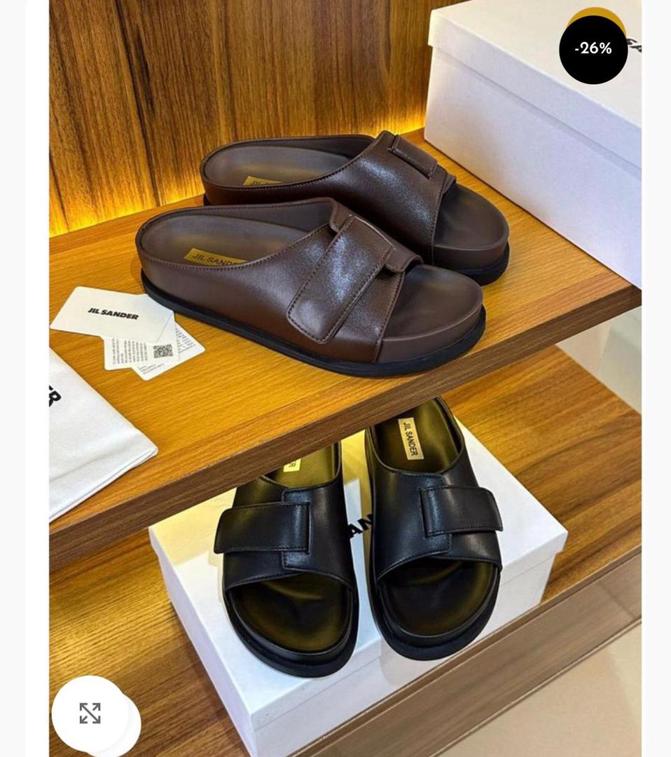 IMG-20260425-WA0025 A pair of brown and black Jil Sander double strap leather slides displayed on a wooden shelf with their respective boxes and packaging.