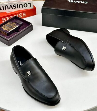 A pair of black leather loafers with interlocking CC logo on a white table, surrounded by luxury brand boxes and books.
