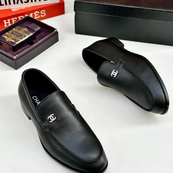 A pair of black leather loafers with interlocking CC logo on a white table, surrounded by luxury brand boxes and books.