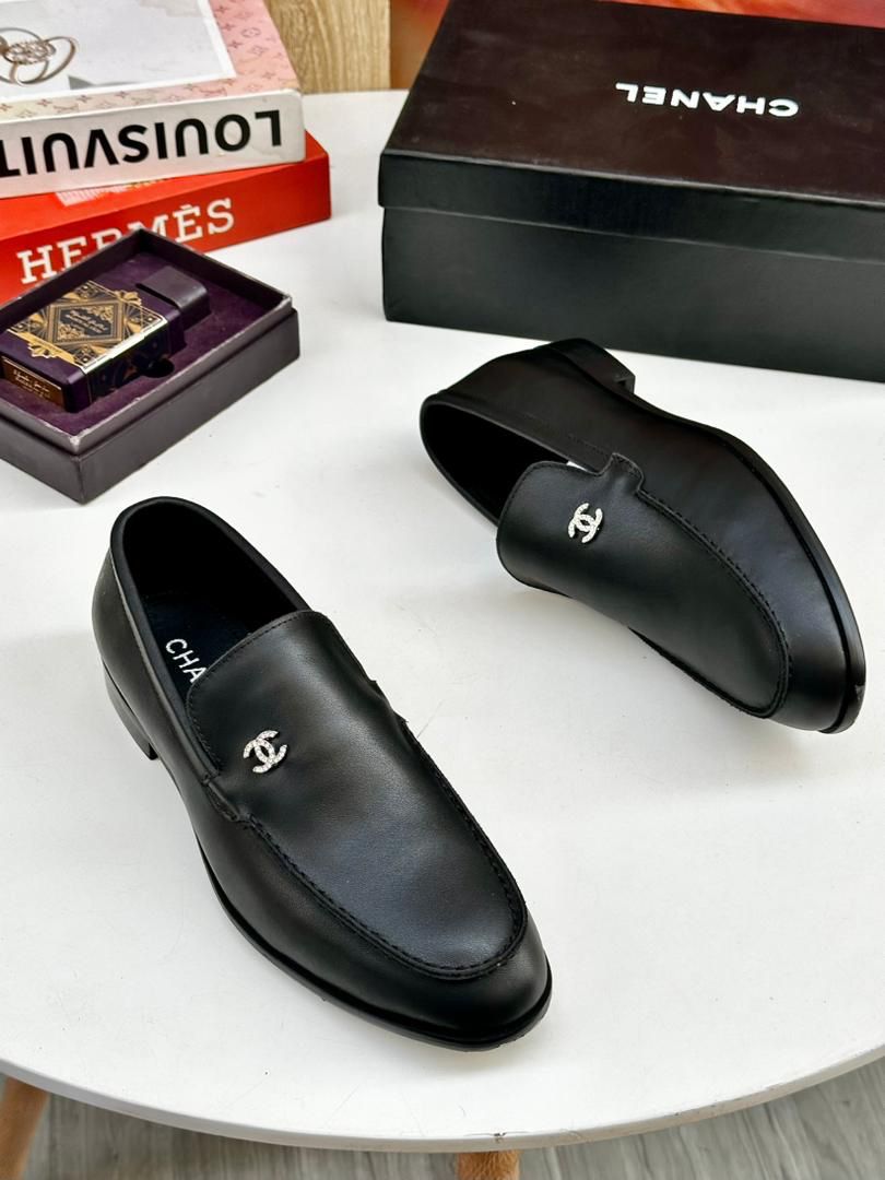 IMG-20260425-WA0027 A pair of black leather loafers with interlocking CC logo on a white table, surrounded by luxury brand boxes and books.