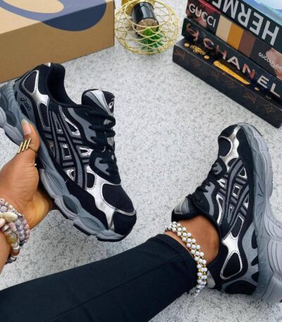 A pair of black and silver luxury sneakers on a woman's feet, with a stylish background featuring luxury brand boxes.
