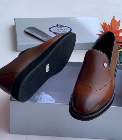 A pair of luxurious brown leather loafers with black soles, displayed in an open gray shoe box with the GENIO logo, accompanied by a shoe tree and a vibrant orange plant.