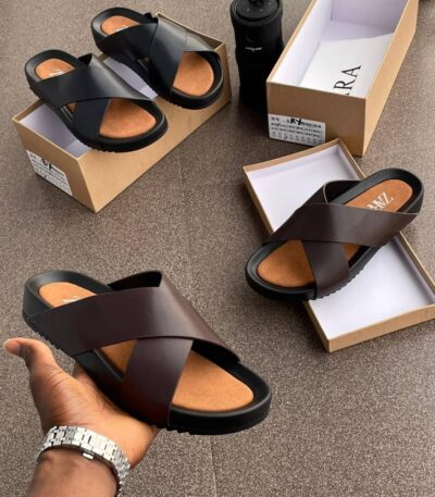 A display of black and brown double strap rubber sandals on a brown surface, with shoe boxes and a hand holding one sandal.