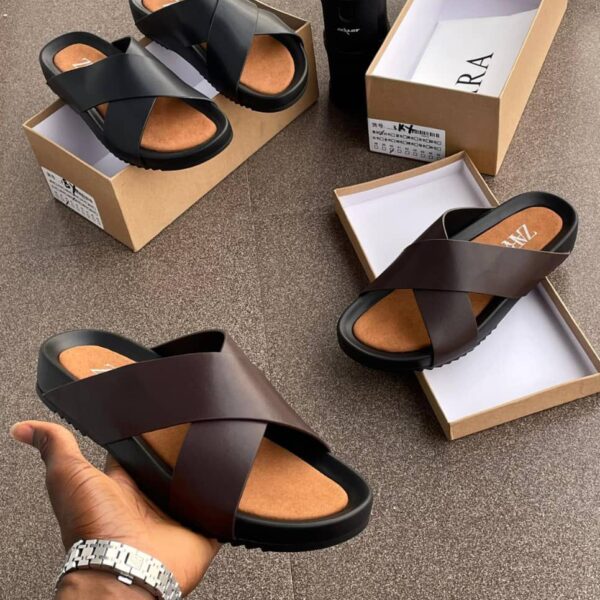 A display of black and brown double strap rubber sandals on a brown surface, with shoe boxes and a hand holding one sandal.