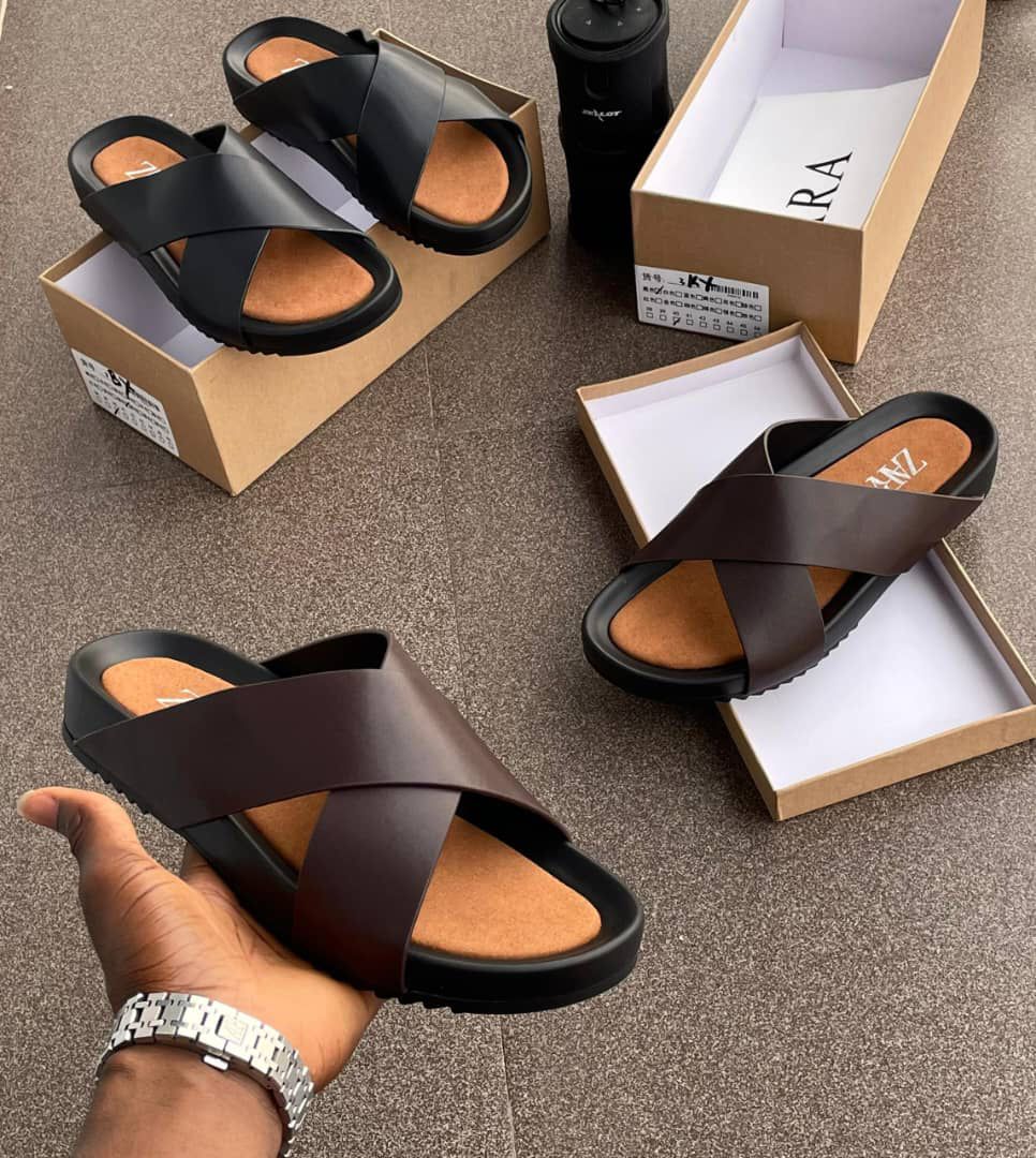 IMG-20260425-WA0031 A display of black and brown double strap rubber sandals on a brown surface, with shoe boxes and a hand holding one sandal.