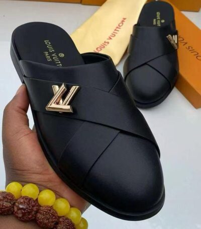 A pair of black leather Louis Vuitton slide sandals with gold-tone logo, presented on a white surface with luxury packaging in the background.