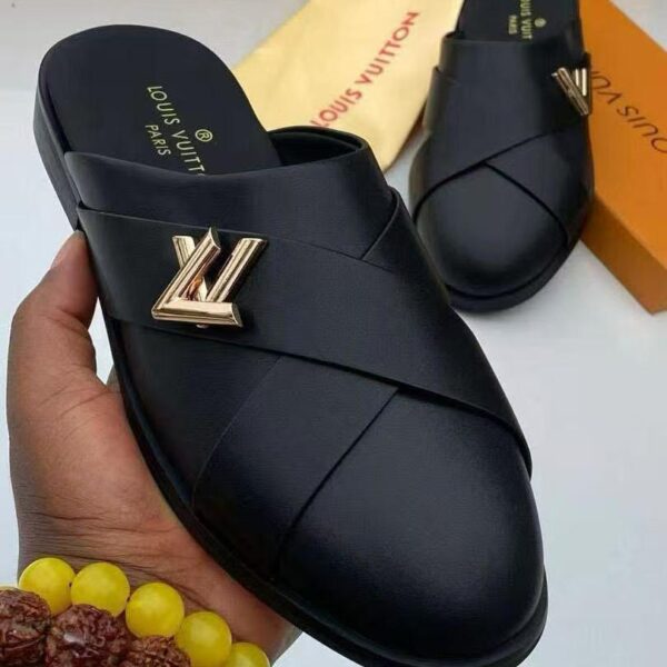 A pair of black leather Louis Vuitton slide sandals with gold-tone logo, presented on a white surface with luxury packaging in the background.