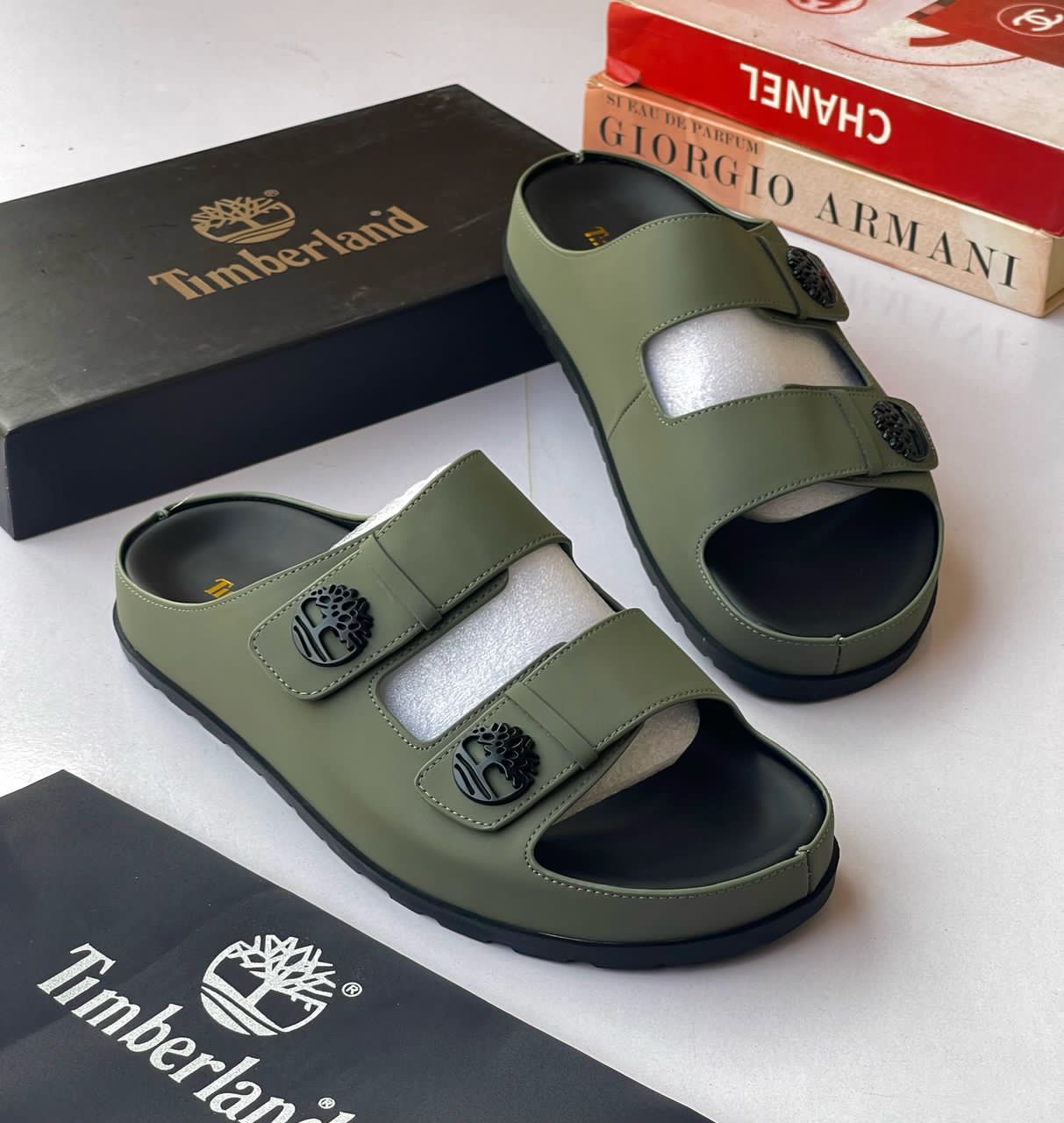 IMG-20260425-WA0035 A pair of olive green Timberland sandals with black soles and logo buckles, displayed with their box and packaging on a white surface.