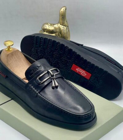A pair of black leather loafers with tassel and buckle detail, displayed on a shoe box with a gold thumbs up statue in the background.