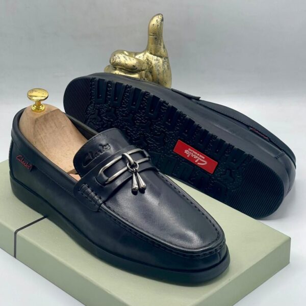 A pair of black leather loafers with tassel and buckle detail, displayed on a shoe box with a gold thumbs up statue in the background.