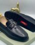 A pair of black leather loafers with tassel and buckle detail, displayed on a shoe box with a gold thumbs up statue in the background.