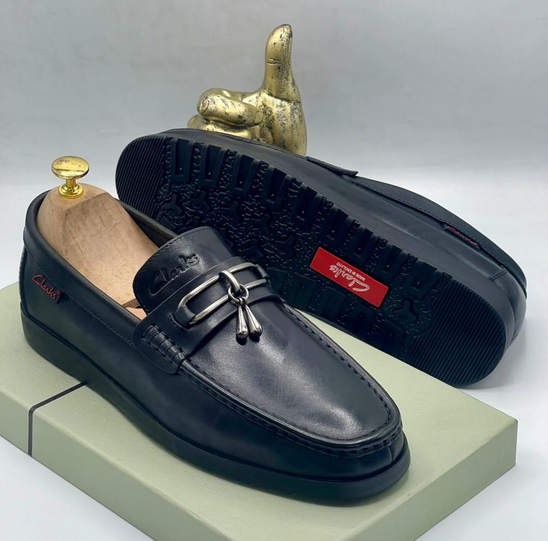 IMG-20260425-WA0036 A pair of black leather loafers with tassel and buckle detail, displayed on a shoe box with a gold thumbs up statue in the background.