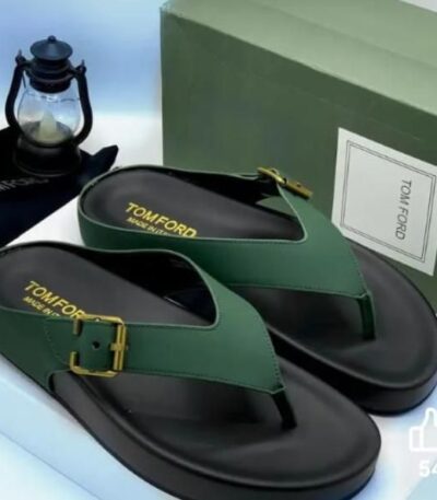 A pair of luxurious green leather thong sandals with gold buckle and TOMFORD branding, displayed on a white surface with a green shoe box and lantern in the background.