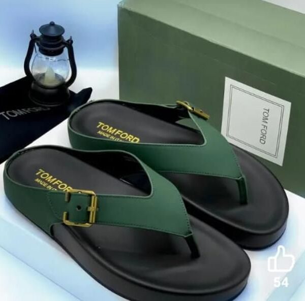 A pair of luxurious green leather thong sandals with gold buckle and TOMFORD branding, displayed on a white surface with a green shoe box and lantern in the background.