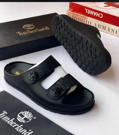 A pair of black leather Timberland slide sandals with two straps and a tree logo buckle, displayed with their box and packaging on a white background.