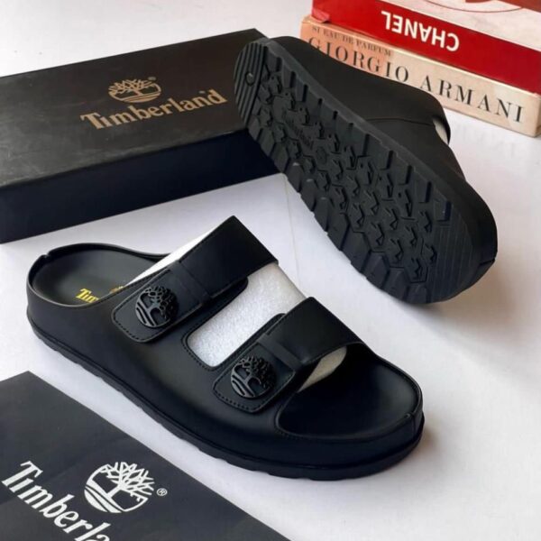 A pair of black leather Timberland slide sandals with two straps and a tree logo buckle, displayed with their box and packaging on a white background.