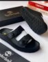A pair of black leather Timberland slide sandals with two straps and a tree logo buckle, displayed with their box and packaging on a white background.