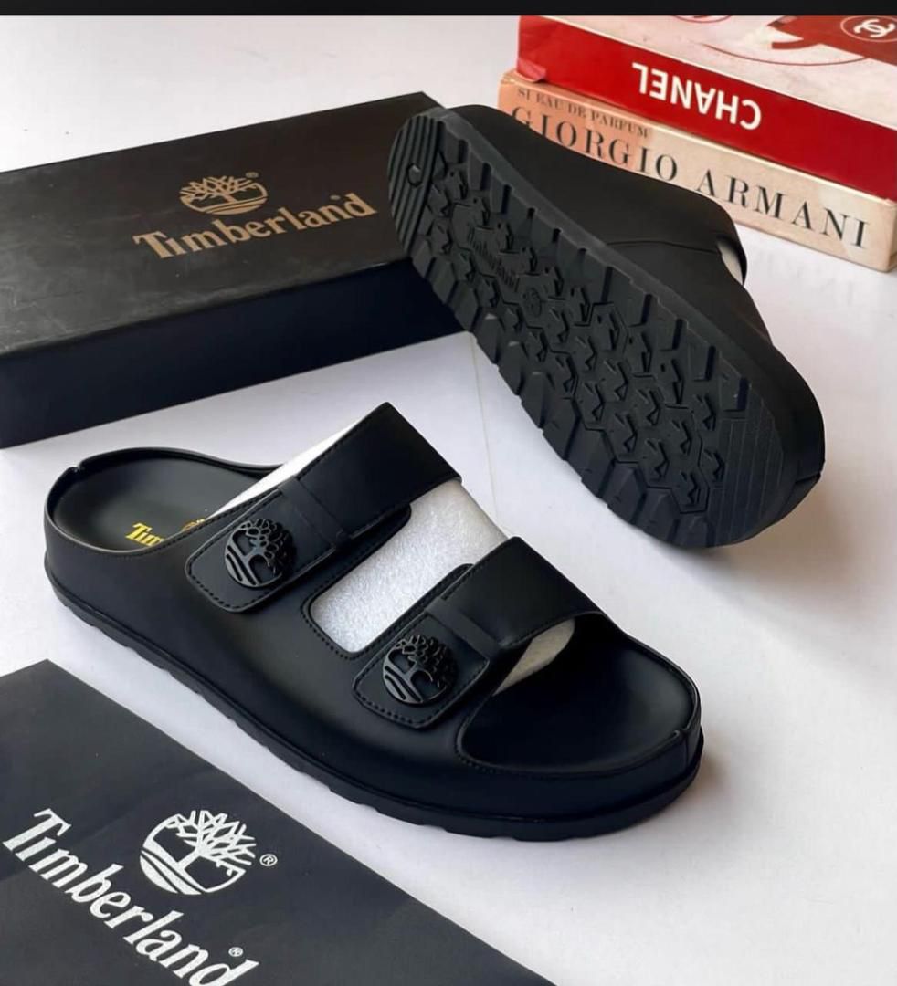 IMG-20260425-WA0038 A pair of black leather Timberland slide sandals with two straps and a tree logo buckle, displayed with their box and packaging on a white background.
