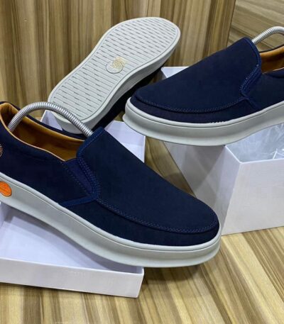 A pair of dark blue suede slip-on shoes with white soles, displayed on shoe boxes, showcasing the Timberland logo and orange accents.