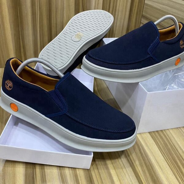 A pair of dark blue suede slip-on shoes with white soles, displayed on shoe boxes, showcasing the Timberland logo and orange accents.