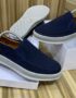 A pair of dark blue suede slip-on shoes with white soles, displayed on shoe boxes, showcasing the Timberland logo and orange accents.