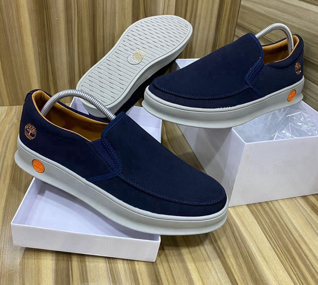 IMG-20260425-WA0044 A pair of dark blue suede slip-on shoes with white soles, displayed on shoe boxes, showcasing the Timberland logo and orange accents.