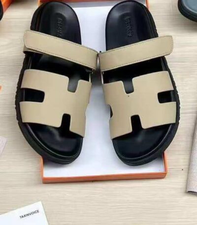 A pair of luxurious black and beige Hermes leather sandals, presented in an open orange box on a light wood surface.